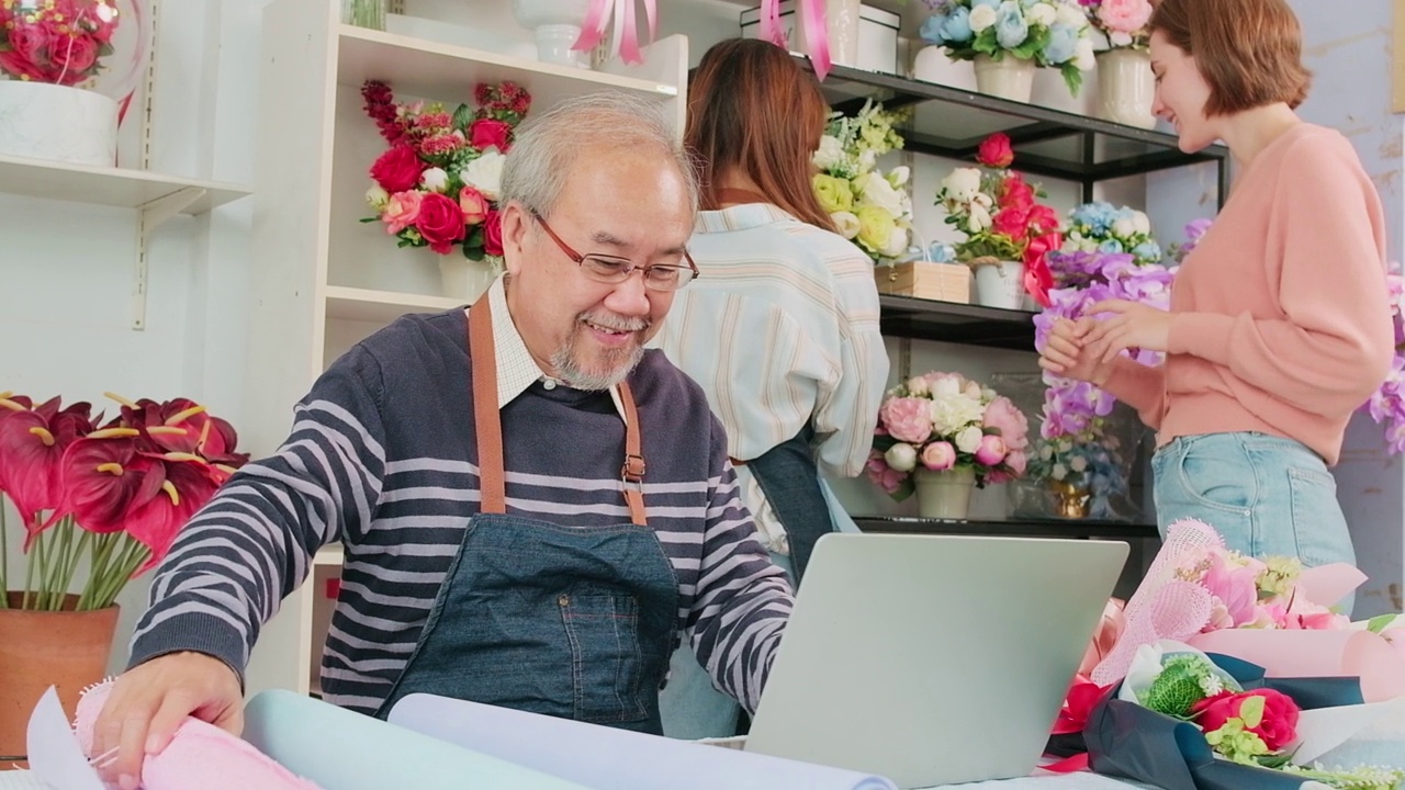 一位年长的亚洲男性老板在一家色彩缤纷的花店工作，手里拿着一台笔记本电脑。视频素材