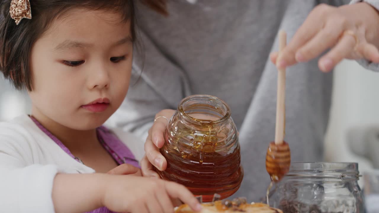 亚洲家庭正在吃早餐，妈妈为小女孩在煎饼上浇蜂蜜，妈妈在家为女儿准备自制的饭菜视频素材