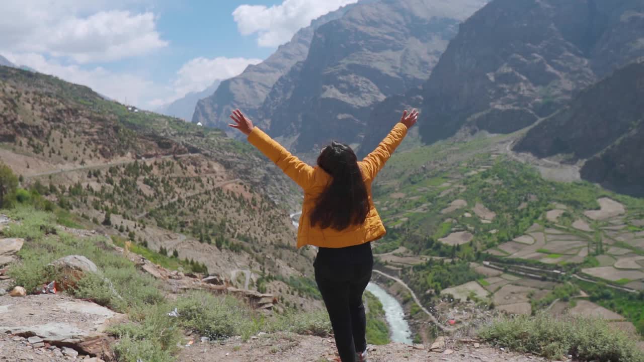 快乐的女徒步旅行者举起手在美丽的山谷和河Bhagha流动的背景在拉豪尔，印度。暑假期间，游客在山上享受视频素材