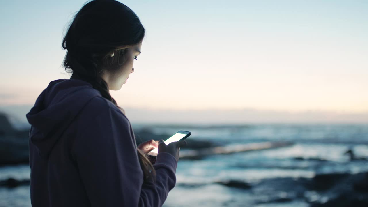 女人，在海滩上用手机和短信模拟交流，社交媒体或在户外浏览。日落时分，女性在海边用智能手机聊天、打字或搜索视频素材