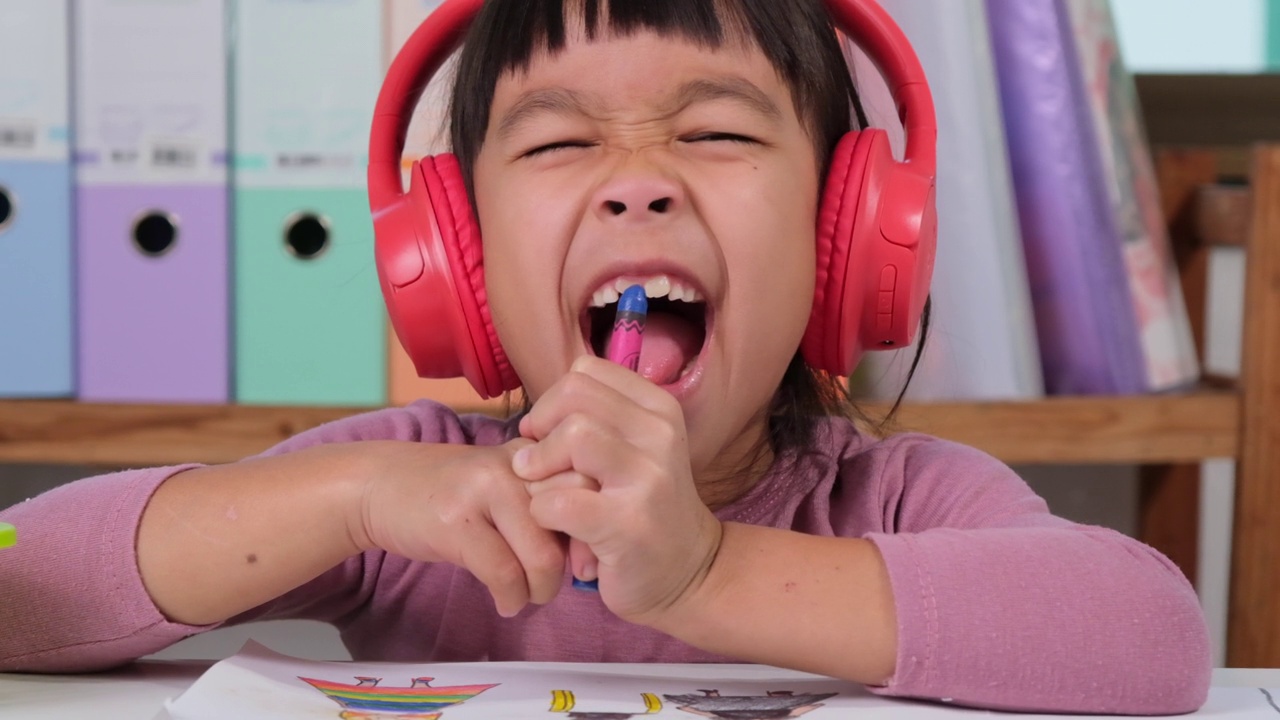 快乐的小女孩戴着耳机，坐在家里房间的桌子前，用彩色铅笔在纸上唱歌和画画。创造力和精细运动技能的发展。视频素材