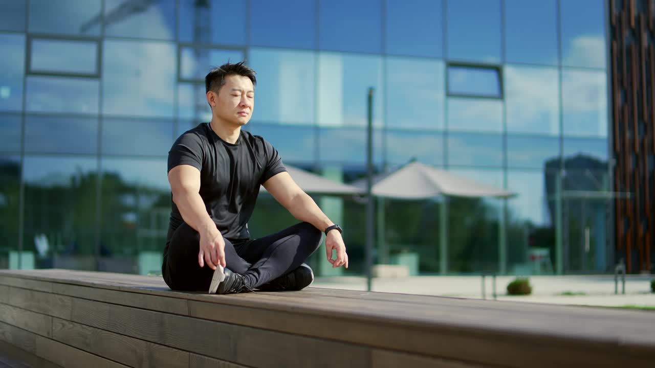 年轻的成年亚洲男子坐在一个现代城市建筑前的长凳上冥想莲花位置。视频素材