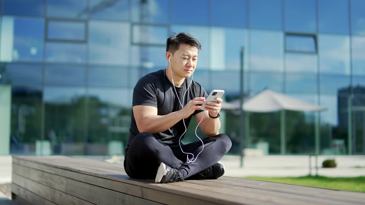 一名成年亚洲男子戴着耳机坐在一座现代城市大楼前的长凳上听音乐。穿着运动服的英俊男子拿着智能手机视频素材