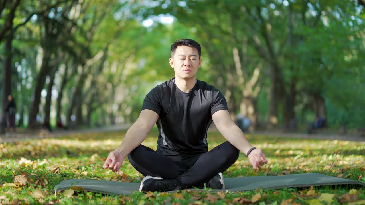 成年亚洲男子在城市公园的席子上冥想莲花位置。穿着运动服的帅哥在外面做瑜伽。视频素材