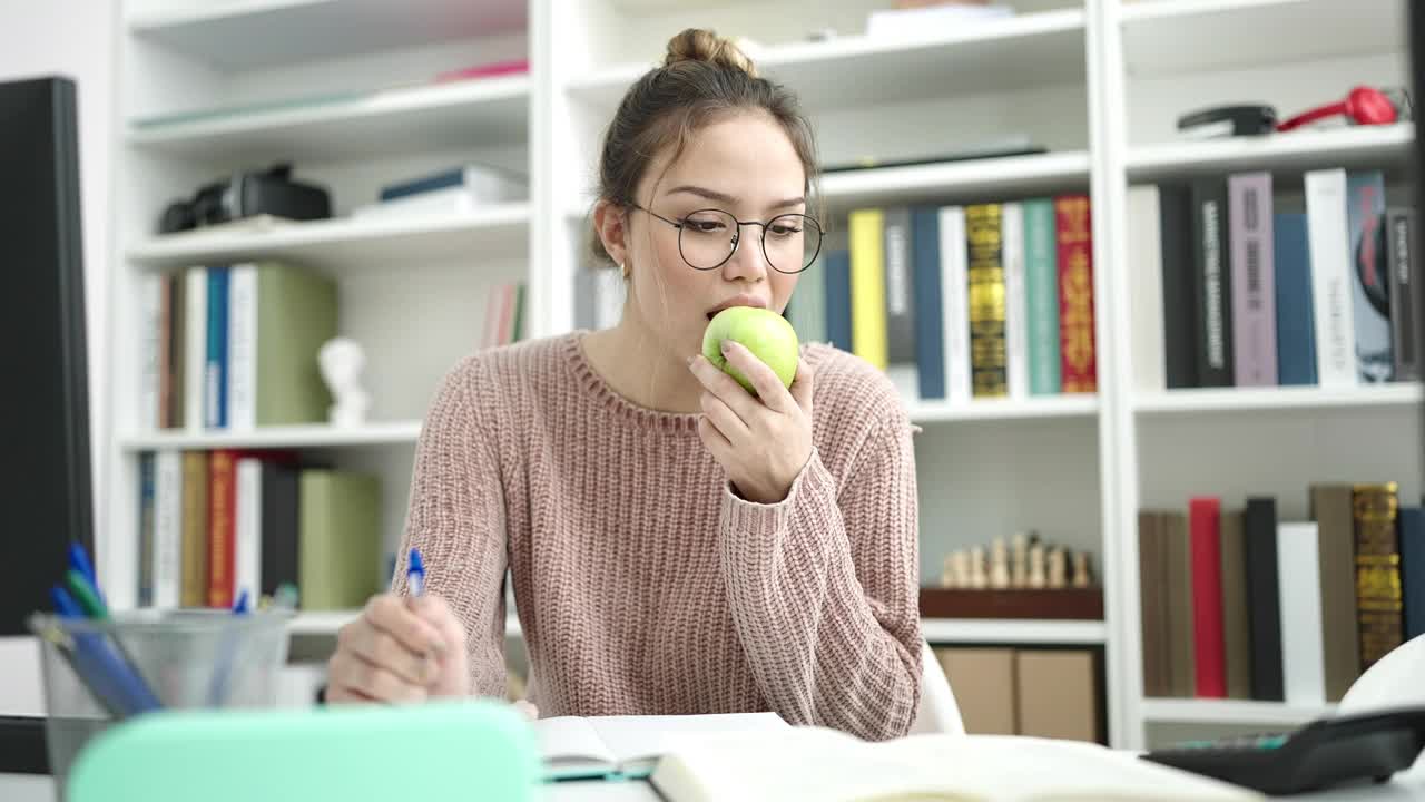 年轻美丽的西班牙女学生写在笔记本上吃苹果在图书馆大学视频素材