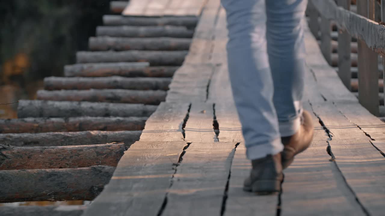 人的腿脚沿着悬索桥在河上。穿着牛仔裤和皮靴的旅行者徒步旅行视频素材