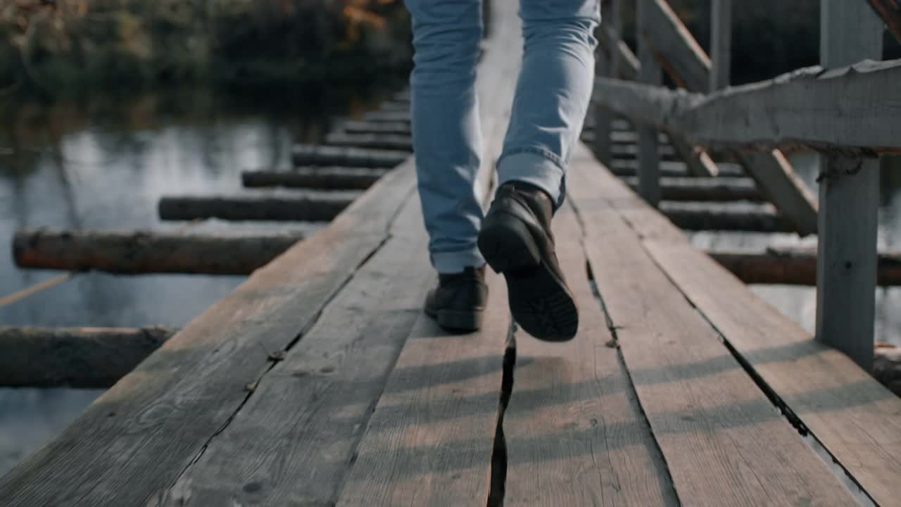 人的腿脚沿着悬索桥在河上。穿着牛仔裤和皮靴的旅行者徒步旅行视频素材