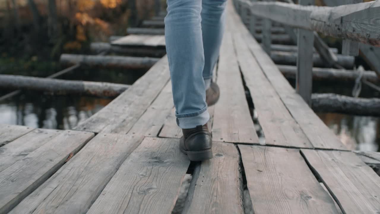 人的腿脚沿着悬索桥在河上。穿着牛仔裤和皮靴的旅行者徒步旅行视频素材