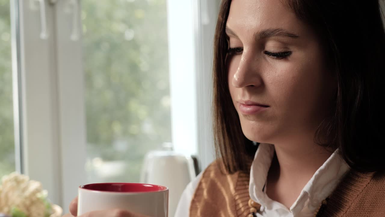 一位女士正在喝咖啡。喝着咖啡或茶的女人享受着味道，看着窗外。特写和慢镜头视频素材