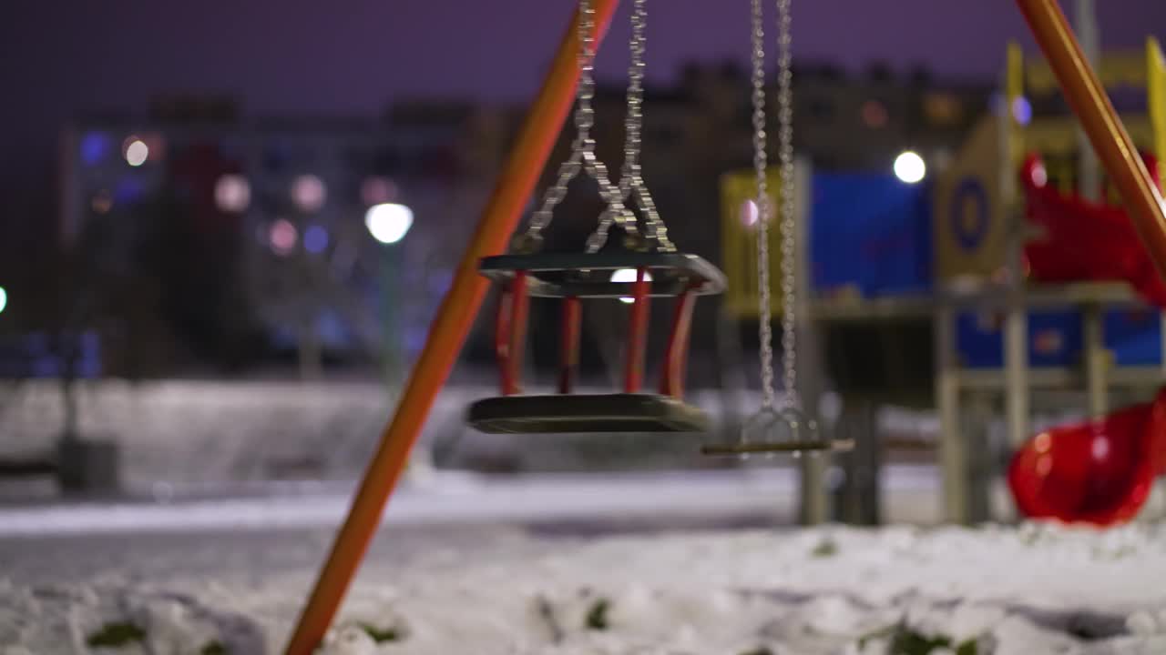 公园里空荡荡的儿童游乐场，孩子的秋千在寒冷的冬夜摇摆，城市背景上有雪视频素材