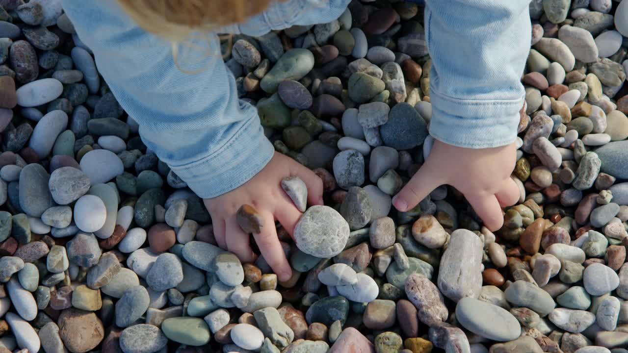 小女孩是不认识的石头海滩上玩和挖在她的手，自上而下的观点，孩子在海滩上玩。视频素材