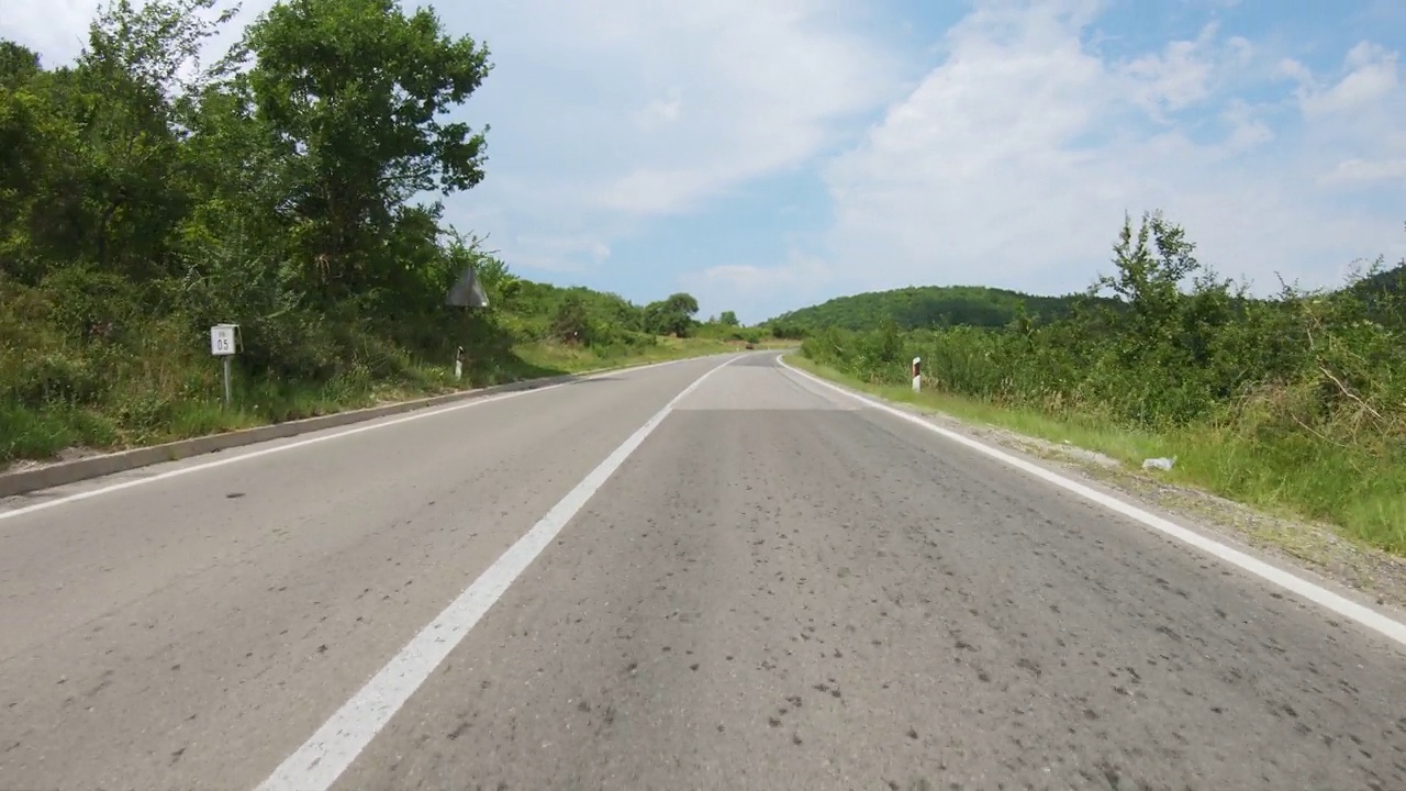 在树木环绕的崎岖曲折的山路上缓慢上坡的汽车或摩托车。山在远方，蓝天有云。观点拍摄视频素材