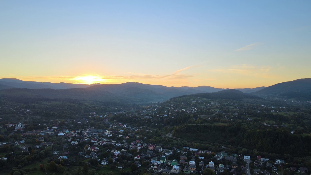 傍晚雾蒙蒙的鸟瞰图，在明亮的夕阳下，在高高的山峰之间的小山村的房子和深色的松林。黄昏时分野山林地的迷人景色视频素材