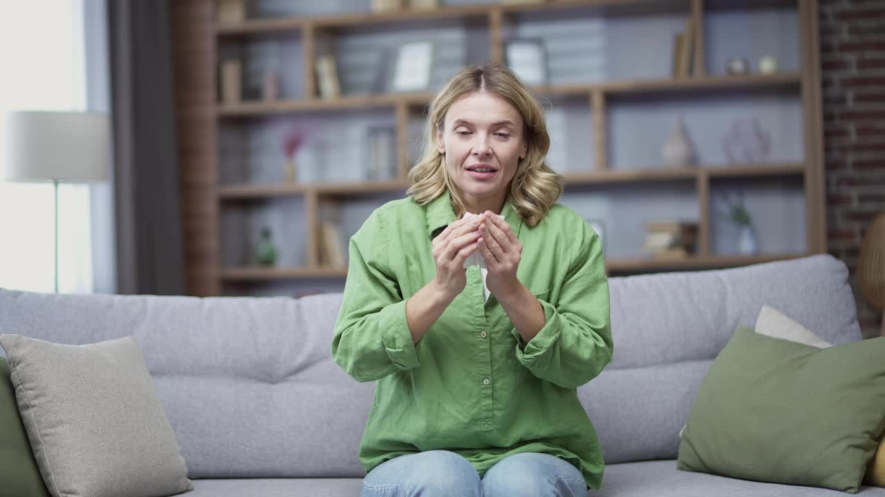 冷成熟的金发女人咳嗽，坐在沙发上在客厅在家里。疲惫的女性穿着便服在室内用手帕擦鼻子视频素材