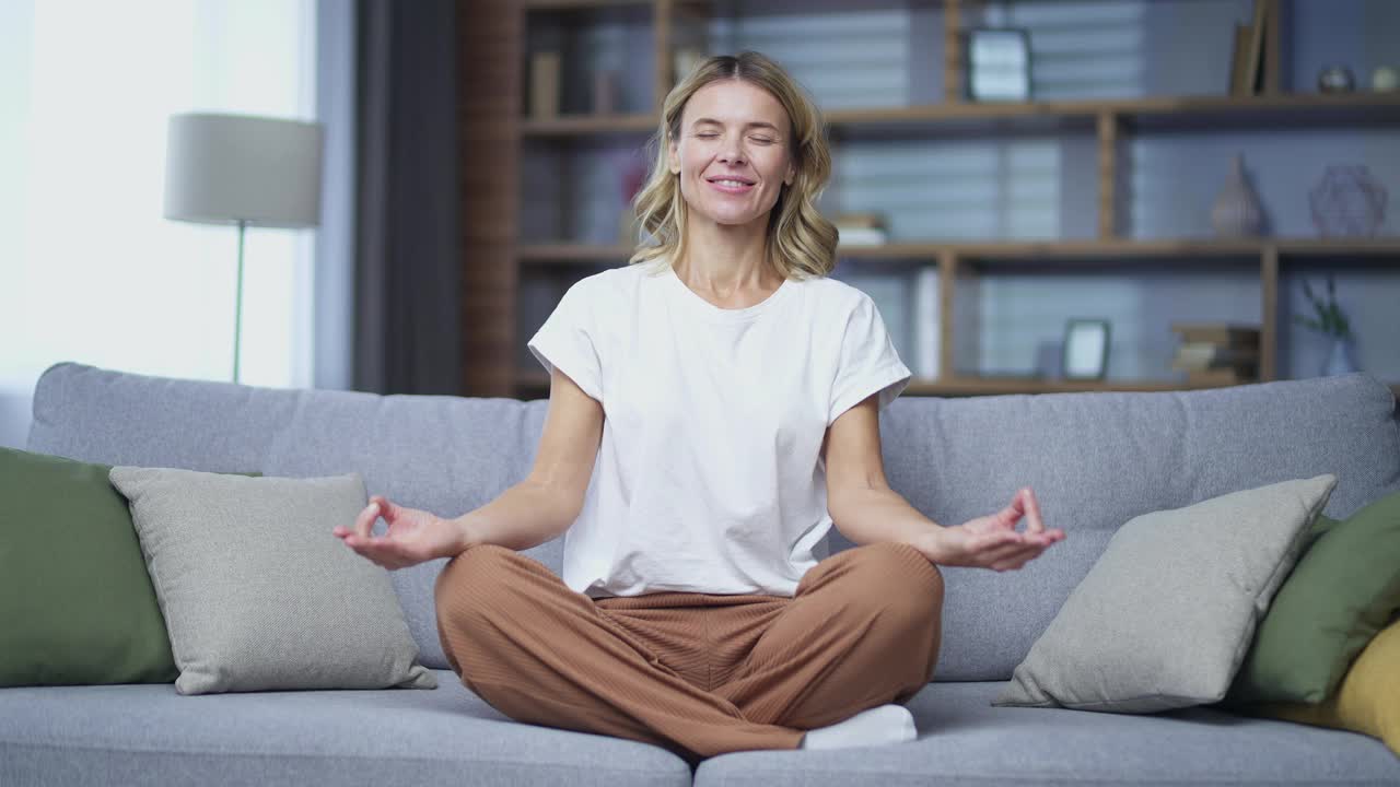 成熟的金发女子闭眼冥想坐在沙发上的莲花位置在客厅在家里。视频素材