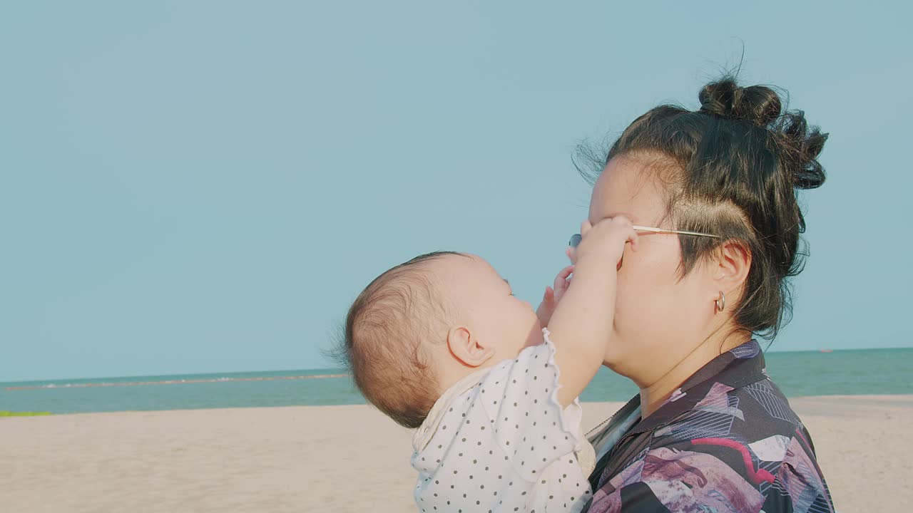 在泰国的夏日假期，一个阳光明媚的日子里，一位亚洲母亲抱着她的小女儿在海边海滩上玩耍视频素材