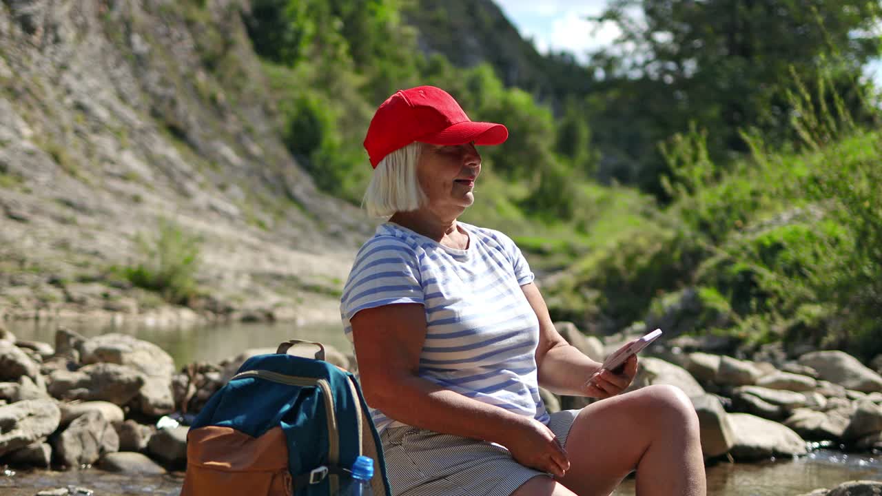 徒步旅行的50多岁女子坐在山河附近的石头上使用智能手机。美丽而鼓舞人心的营地位置，户外氛围，生活方式。旅游的概念视频素材