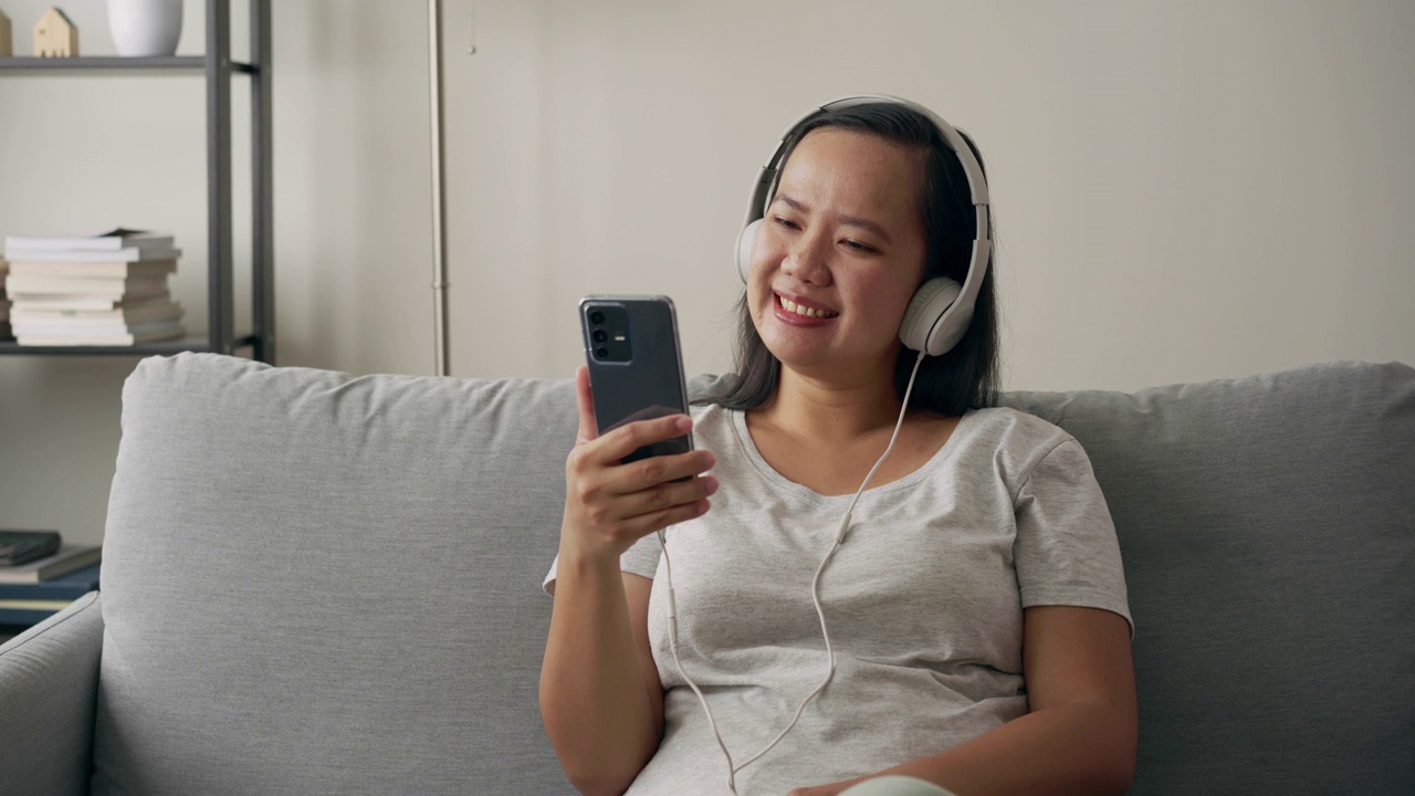 快乐的女人戴着耳机坐在沙发上，在家里放松，喜欢用智能手机听音乐视频素材