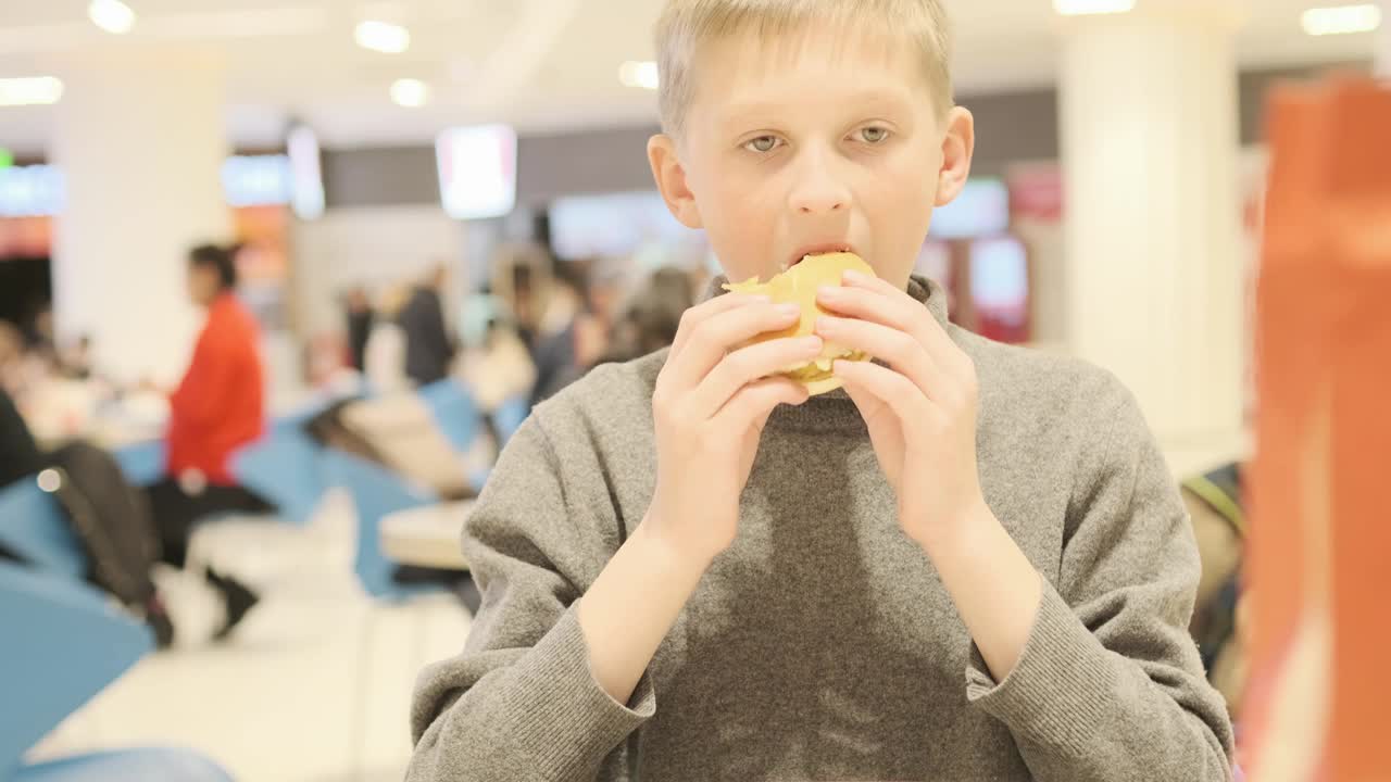 十几岁的男孩在吃汉堡。饥饿的孩子在快餐店。饥饿的孩子在餐厅吃汉堡。快餐店。有害的营养。小男孩坐在咖啡馆里吃起司汉堡视频素材