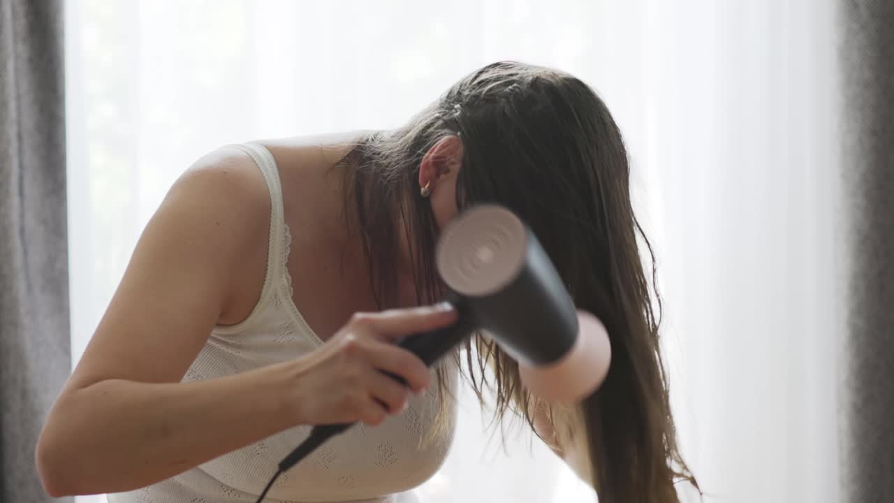 漂亮的年轻女子使用吹风机附近的窗户在家里视频素材