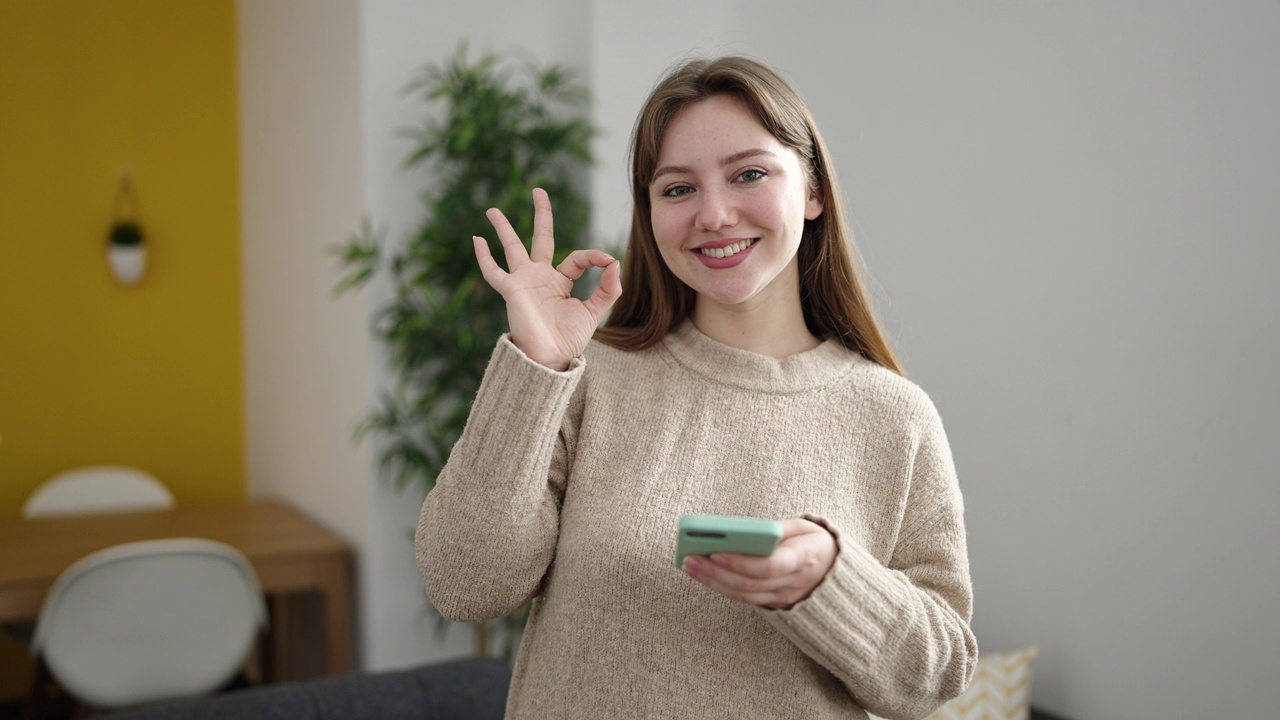 年轻的金发女子使用智能手机在家里做ok的手势视频素材