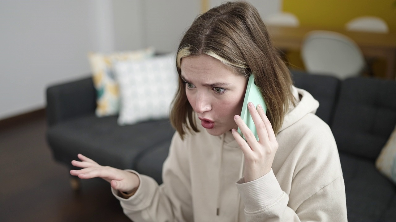 年轻的金发女人在家里用智能手机说话，表情不开心视频素材