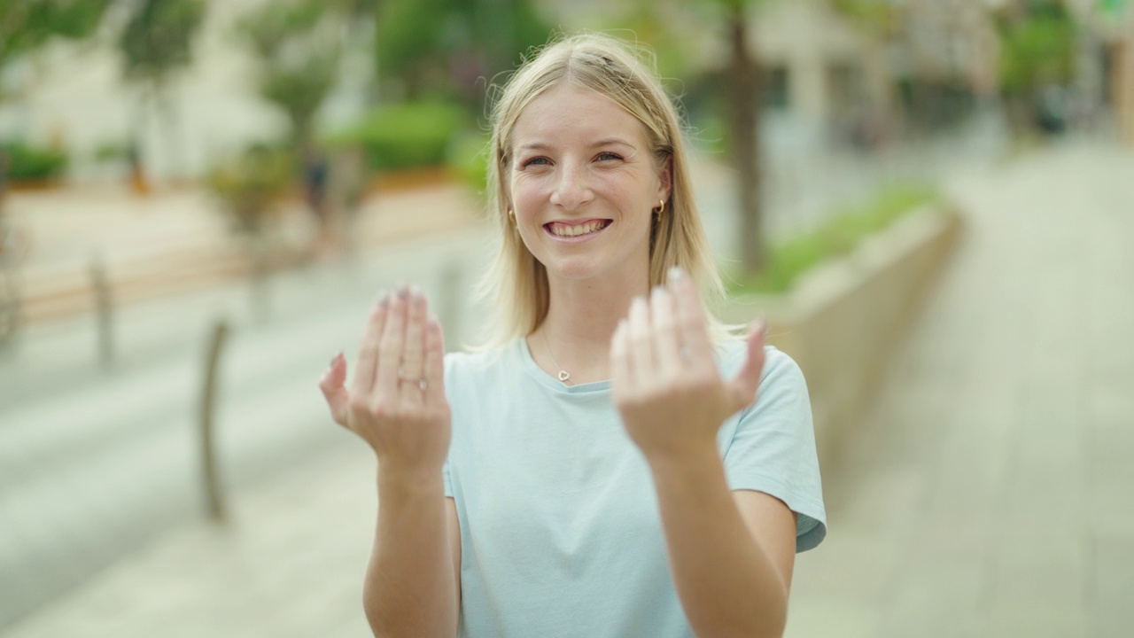 年轻的金发女人微笑自信做即将到来的手势与手在街上视频素材