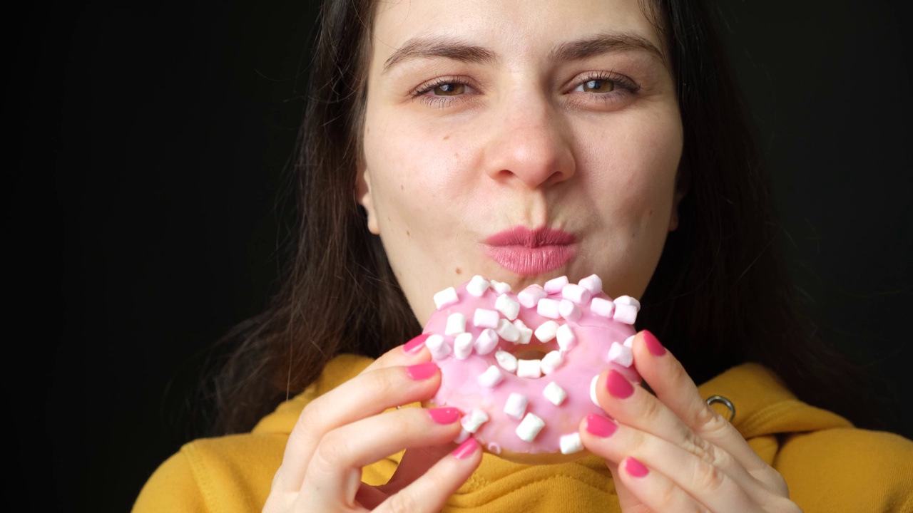 一个女人看着粉红色的棉花糖甜甜圈舔了舔，然后咬了一口，黑色的背景。视频素材