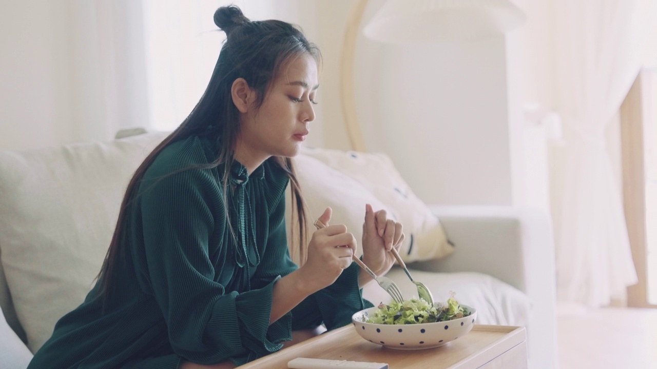 年轻快乐的亚洲女人坐在客厅的沙发上吃健康的沙拉。视频素材