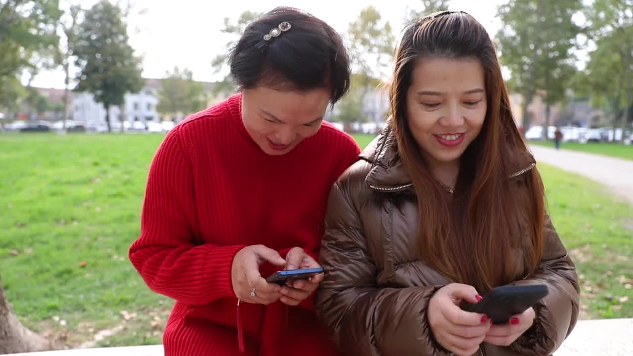 两名中国女子坐在公园的长椅上玩智能手机视频素材