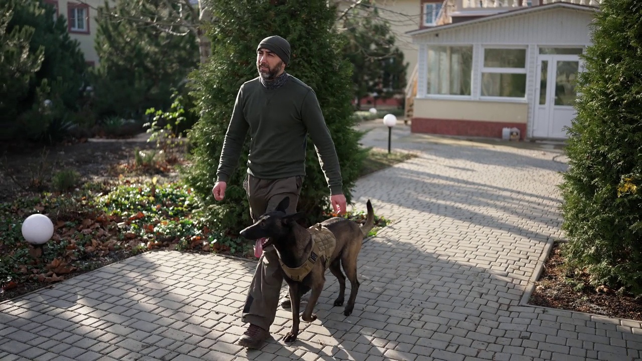 一名男子带着戴着军用防护项圈的服务犬走过房子的院子视频素材