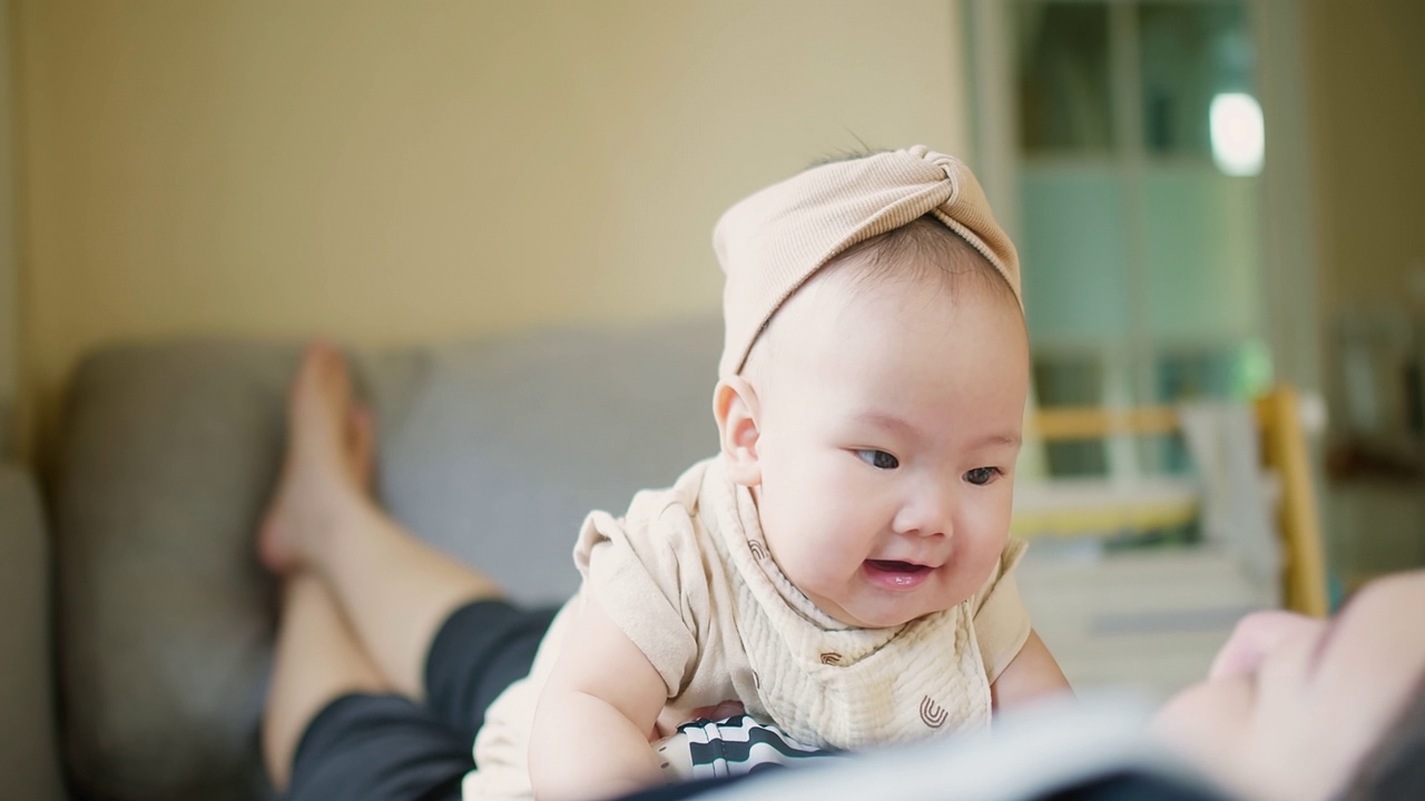 快乐的妈妈和宝宝一起在客厅玩亲密活动。小可爱的亚洲婴儿婴儿女孩兴奋的笑和有趣的花时间在沙发上的家庭在家里玩得很开心视频素材