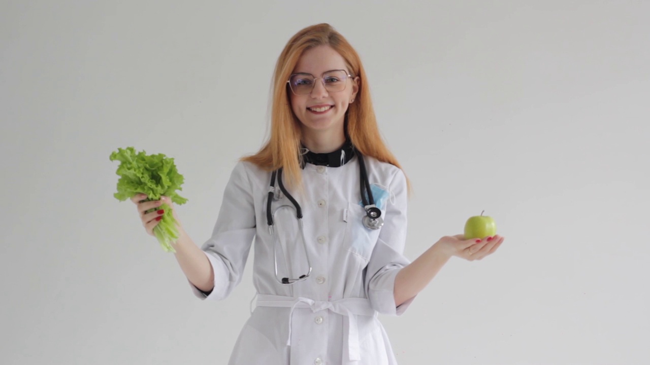 一个红头发的年轻女医生手里拿着苹果和青菜，在一个孤立的白色背景上提出了健康和适当食物的概念。视频素材