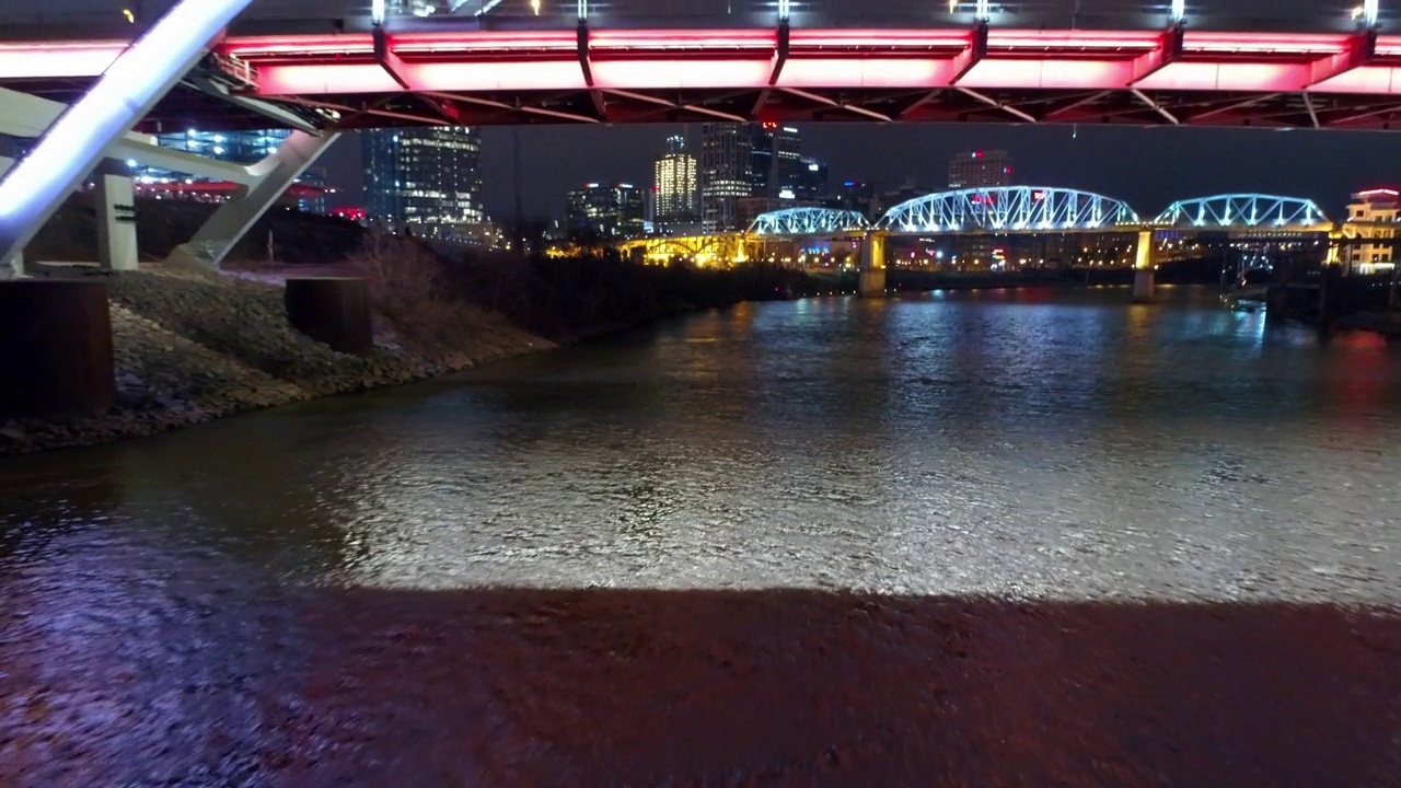 航拍:现代城市中被照亮的著名桥梁，无人机在夜晚向后飞越河流-田纳西州纳什维尔视频素材