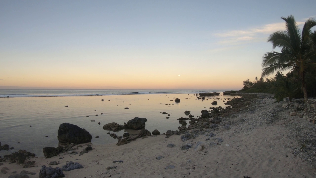 手持静止的海洋海岸线海湾沙质岩石海滩与椰子树在日落月亮升起时，在泻湖和平蛤水波内与晴朗的天空眺望东太平洋的地平线视频素材