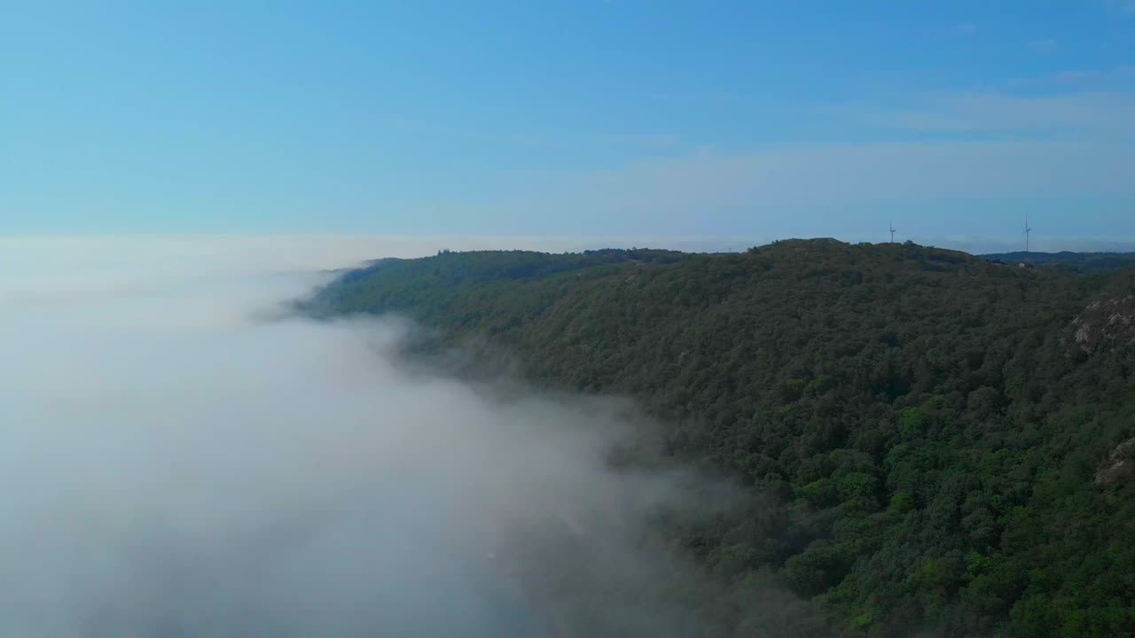 倾斜向下，俯瞰着雾气弥漫的山谷，山谷里有木制的小山。远处有两个风力涡轮机。视频素材