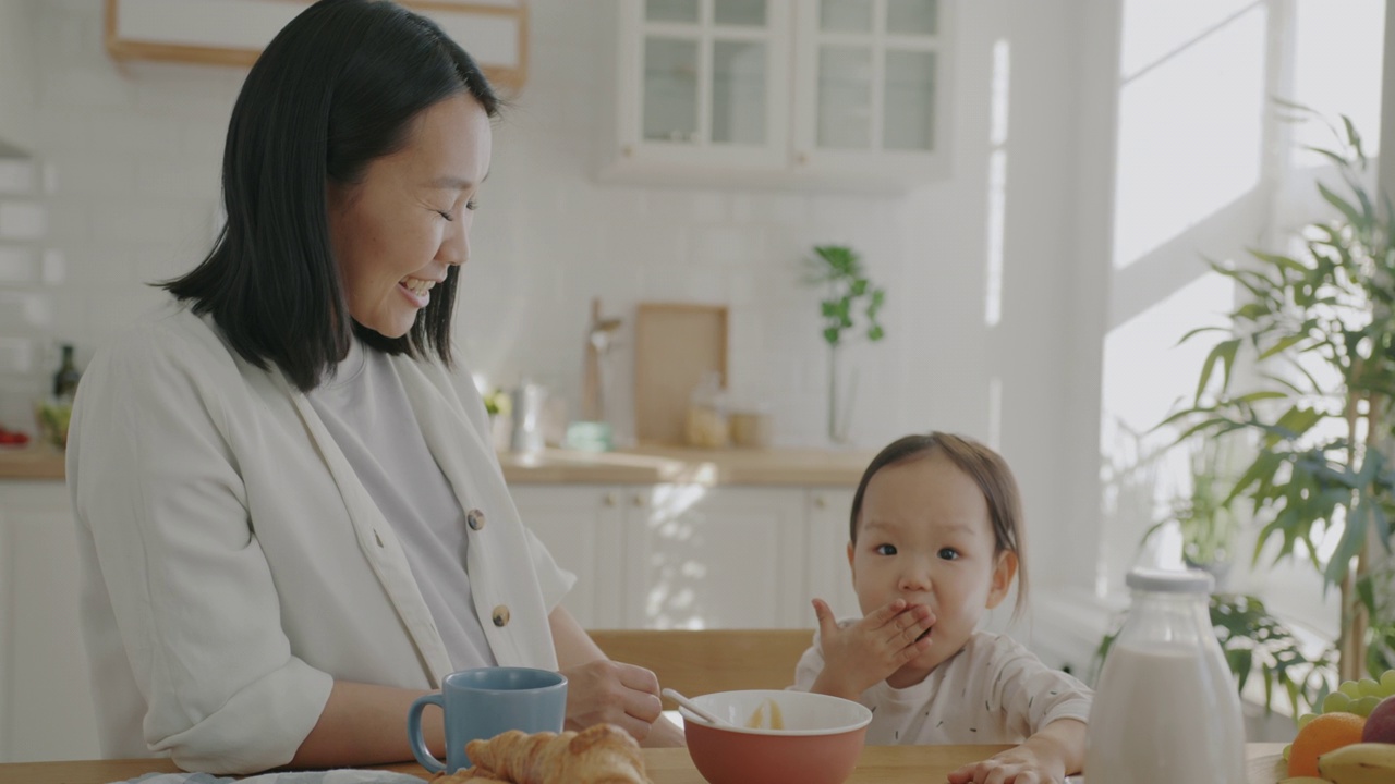 年轻的亚洲女人在家里的厨房里喂可爱的小孩，笑视频素材