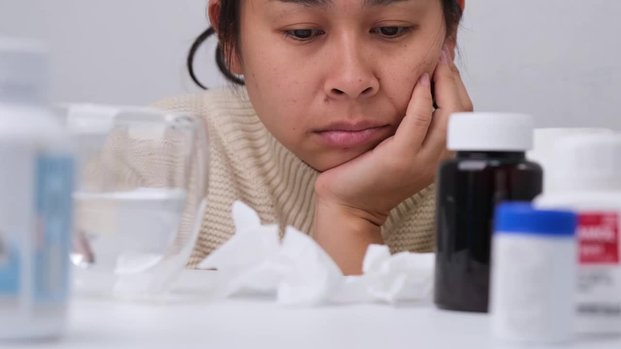 亚洲年轻女子坐在那里看着很多药片，感觉不舒服。亚洲女性因为不想吃药而感到压力和无聊。视频素材