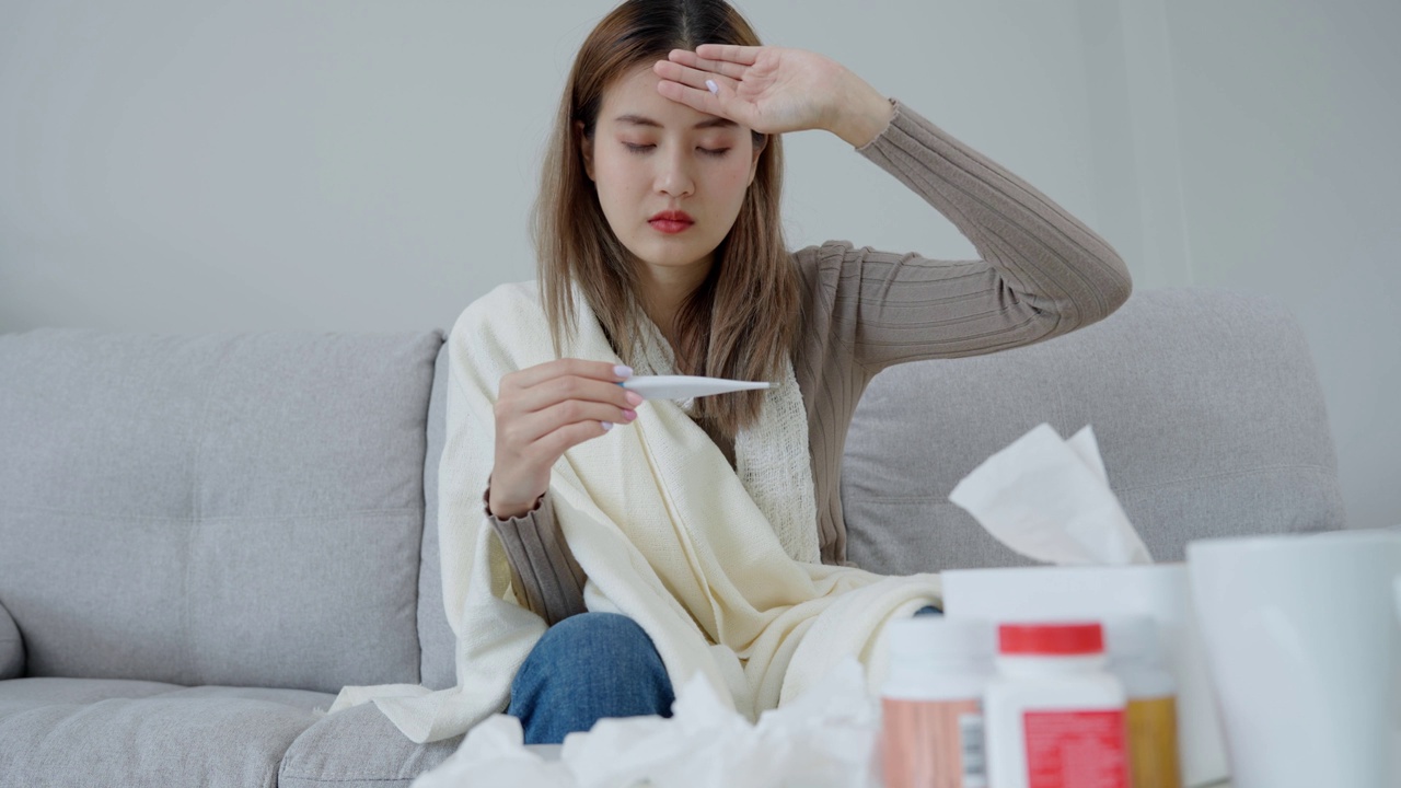 年轻的亚洲女性高烧，同时检查体温，女性打喷嚏和流鼻涕与季节性流感，过敏，数字体温计，病毒，冠状病毒，疾病，呼吸道视频素材