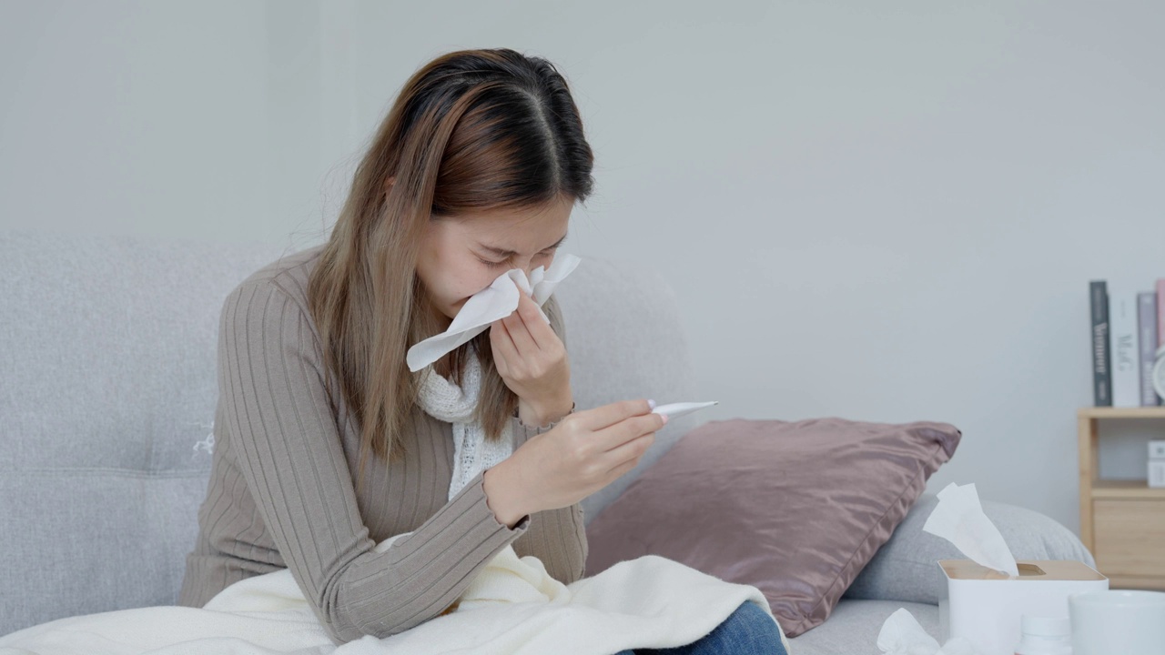 年轻的亚洲女性高烧，同时检查体温，女性打喷嚏和流鼻涕与季节性流感，过敏，数字体温计，病毒，冠状病毒，疾病，呼吸道视频素材