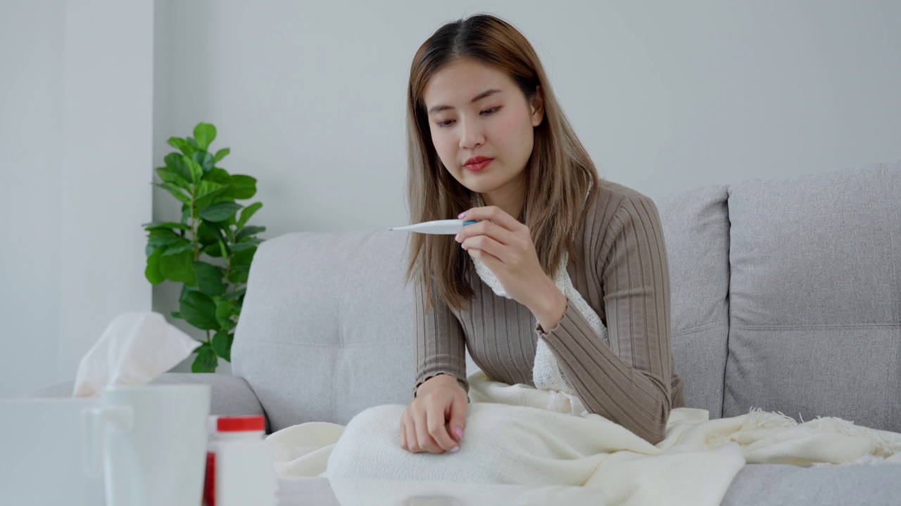 年轻的亚洲女性高烧，同时检查体温，女性打喷嚏和流鼻涕与季节性流感，过敏，数字体温计，病毒，冠状病毒，疾病，呼吸道视频素材