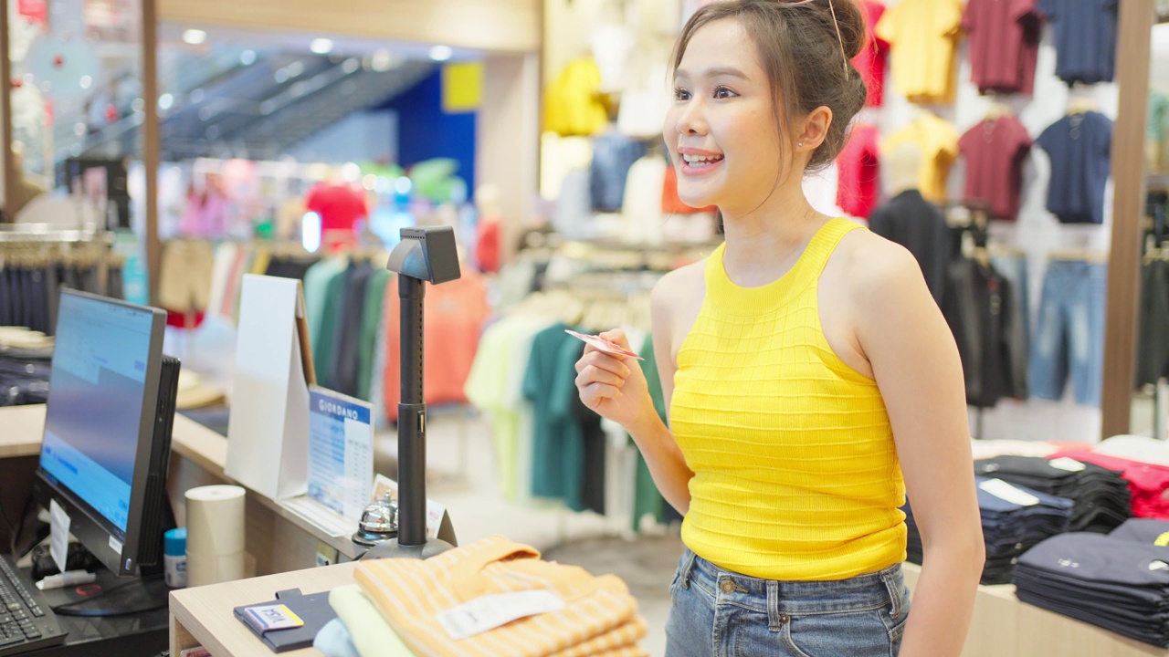 亚洲年轻女性在购物中心使用信用卡支付服装产品。迷人的快乐女孩喜欢在商场中心的衣架上走在百货商店里选择衣服和新t恤。视频素材