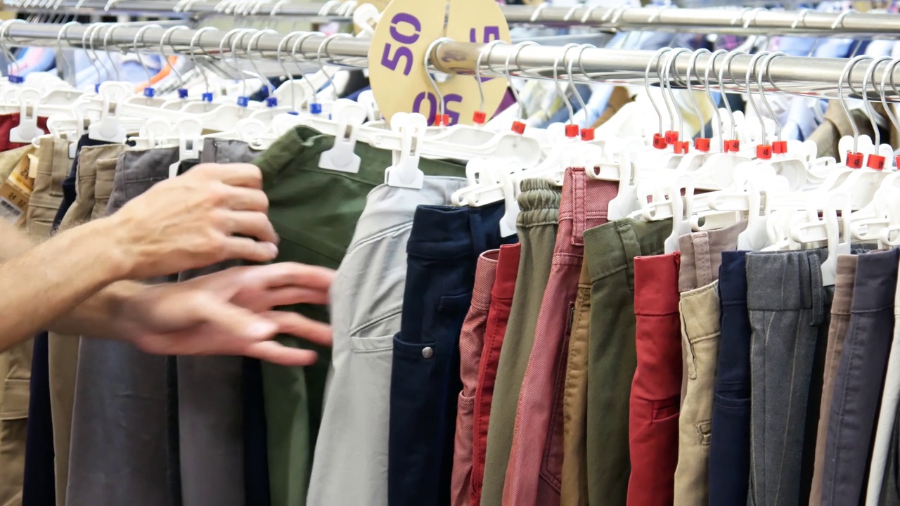 服装店货架上挂着许多裤子的特写，一位男性买家选择了一条视频素材