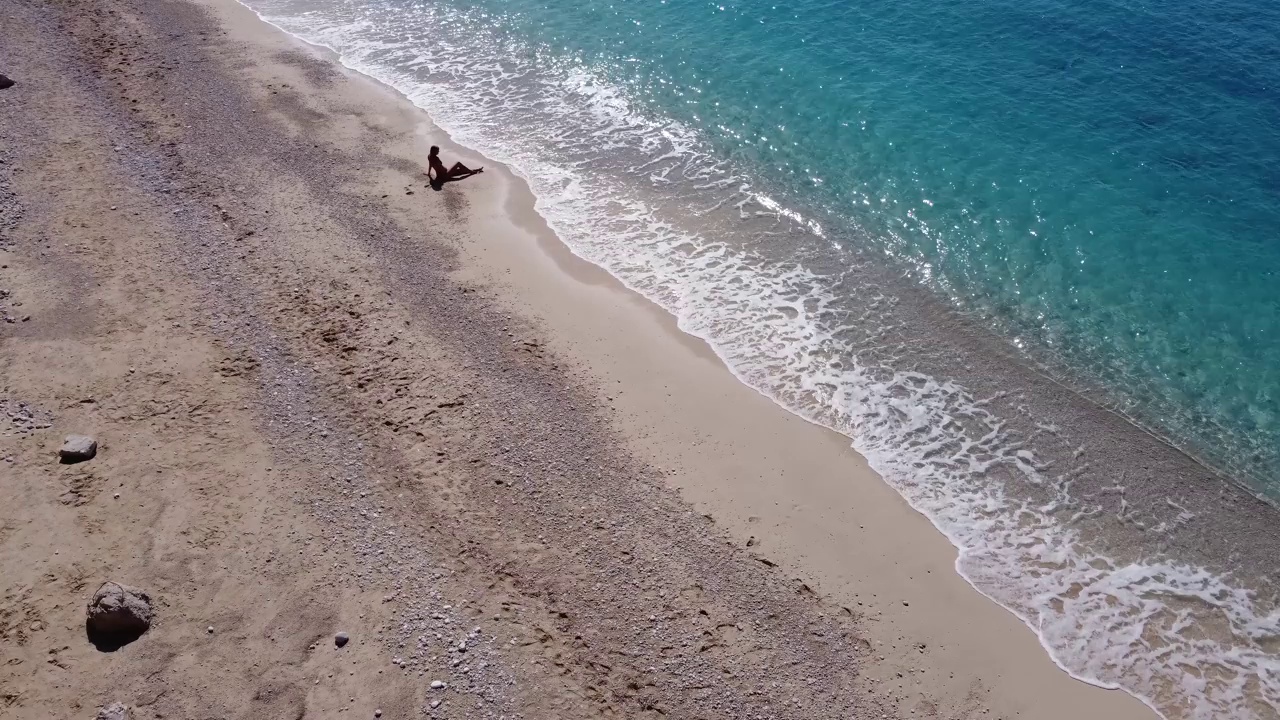 鸟瞰美丽快乐的女子穿着泳装坐在浅海的沙滩上，享受海滩和柔软的蓝绿色海浪。Lefkada岛的Egremni海滩夏季的热带海。视频下载