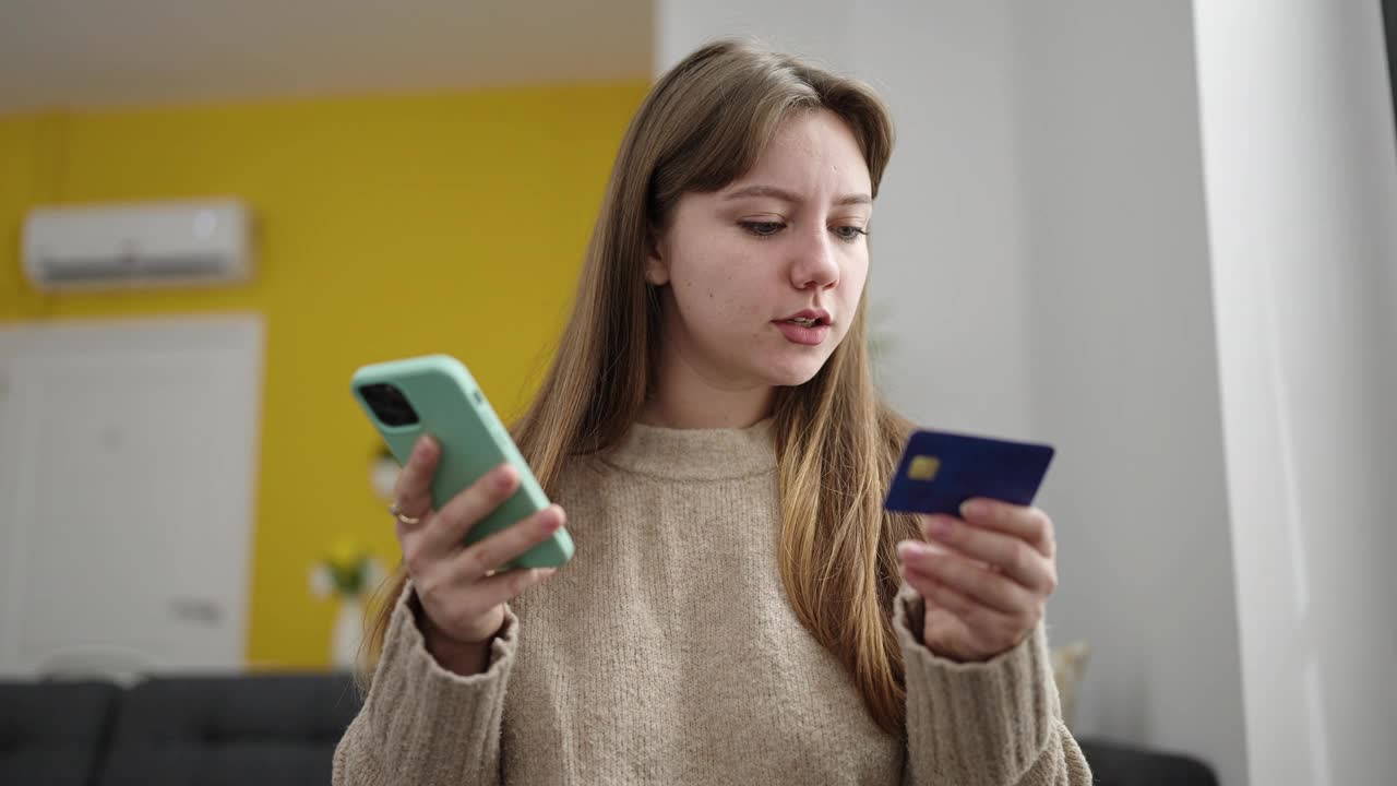 年轻的金发女子使用智能手机和信用卡坐在家里的地板上视频素材