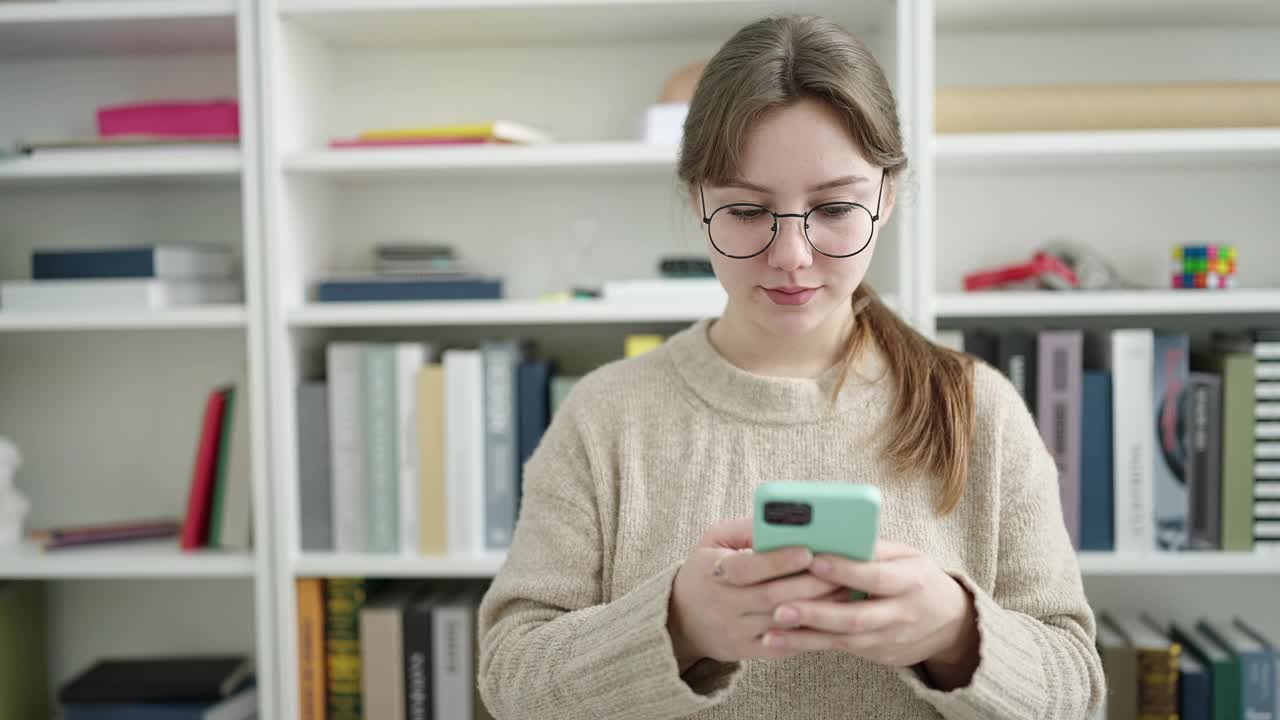 年轻的金发女学生使用智能手机做ok手势在图书馆大学视频素材