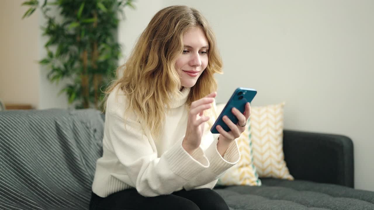 年轻的金发女子坐在家里的沙发上使用智能手机视频素材