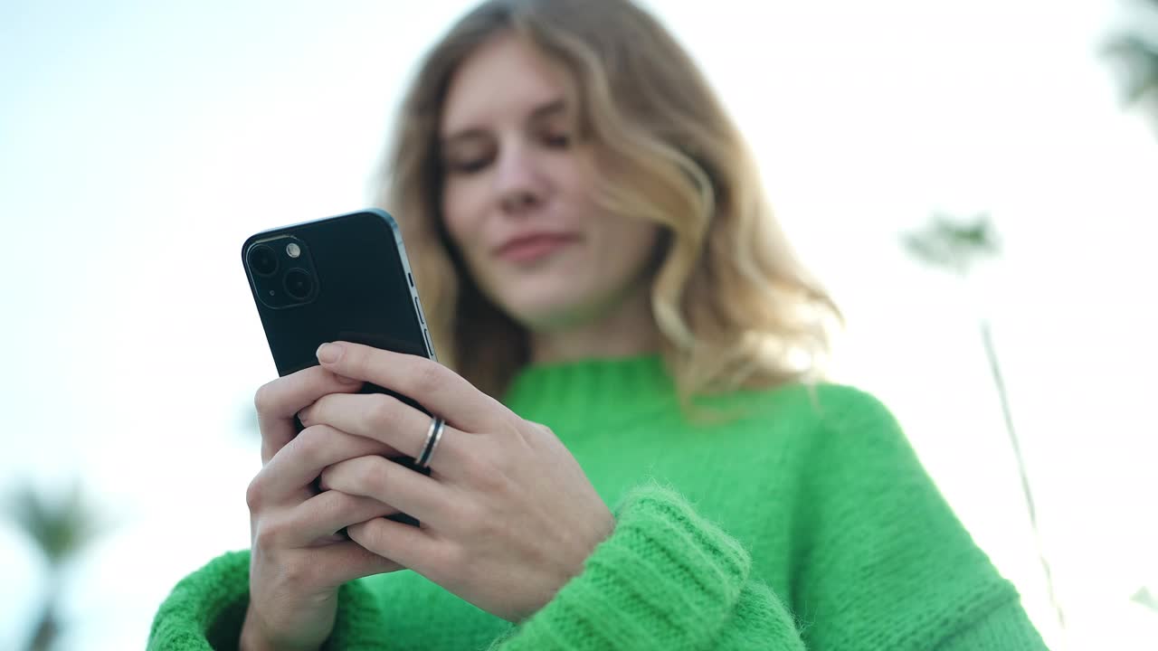 年轻的金发女子在街上自信地微笑着使用智能手机视频素材