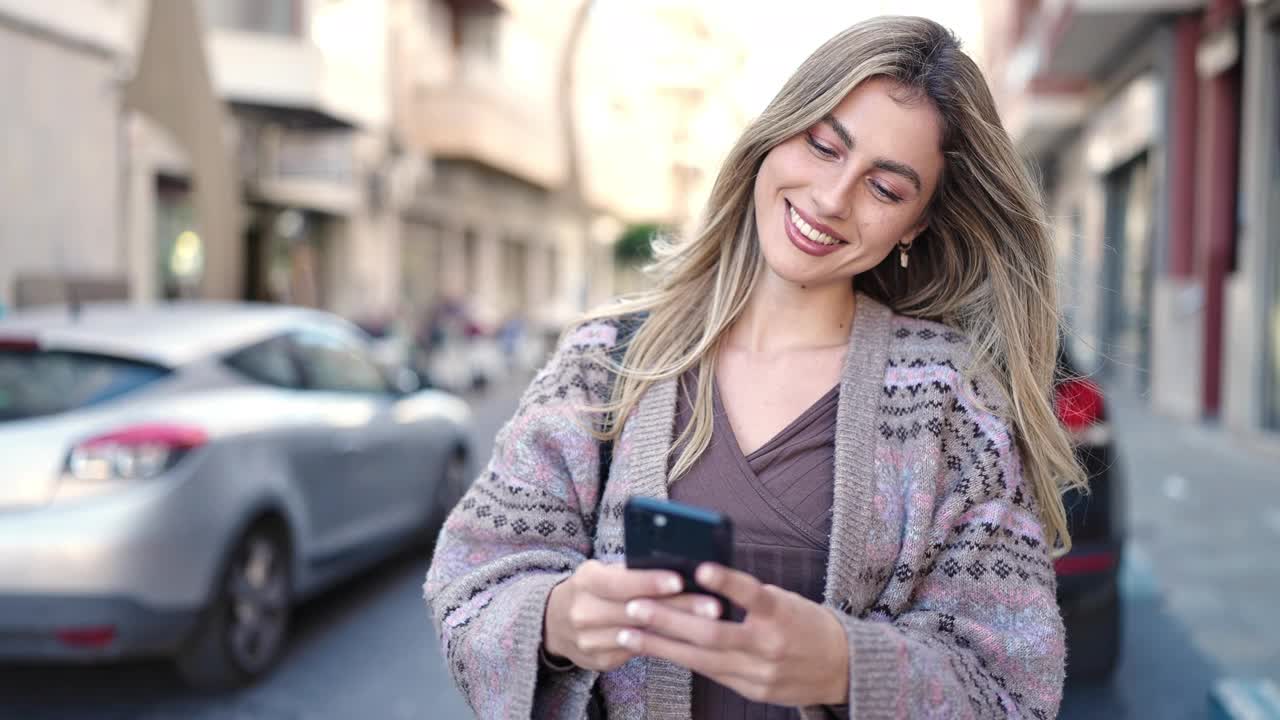 年轻的金发女子在街上自信地微笑着使用智能手机视频素材