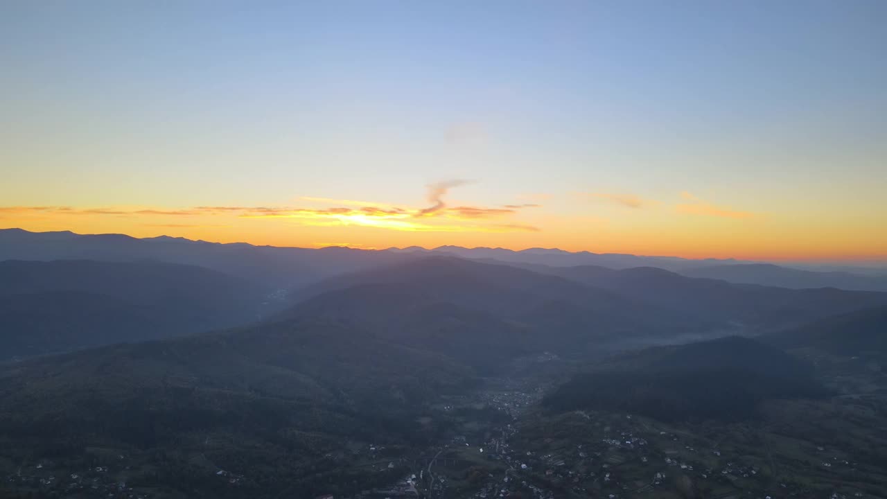 傍晚雾蒙蒙的鸟瞰图，在明亮的夕阳下，在高高的山峰之间的小山村的房子和深色的松林。黄昏时分野山林地的迷人景色视频素材